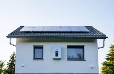 "Modern house with rooftop solar panels and wall-mounted battery storage unit with green trees and cloudy sky in the background."