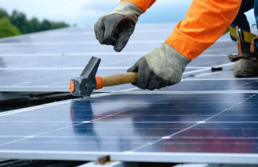 Close-up of a gloved solar installer tapping a solar panel frame into a roof racking clip with a rubber mallet under bright overcast light, with shingles and a safety harness line softly blurred in the background.