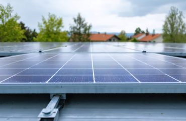Close-up of residential rooftop solar panels on raised standoff mounts showing the airflow gap beneath, under soft overcast daylight with blurred trees and rooflines behind.