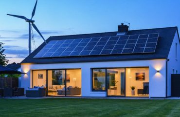 Modern house at twilight with rooftop solar panels and a small residential wind turbine spinning in the yard, warm interior lights visible, with trees and distant hills in the background.